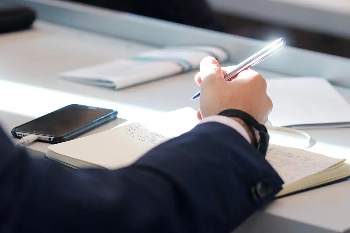 Mână care scrie într-un carnet lângă un telefon, pe o masă de lucru