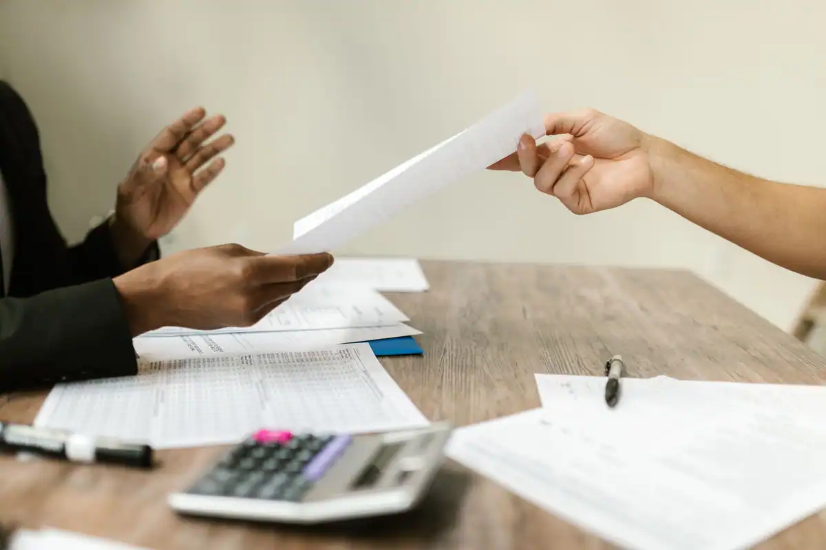 Mâini care își dau un document peste masă, lângă calculator și foi