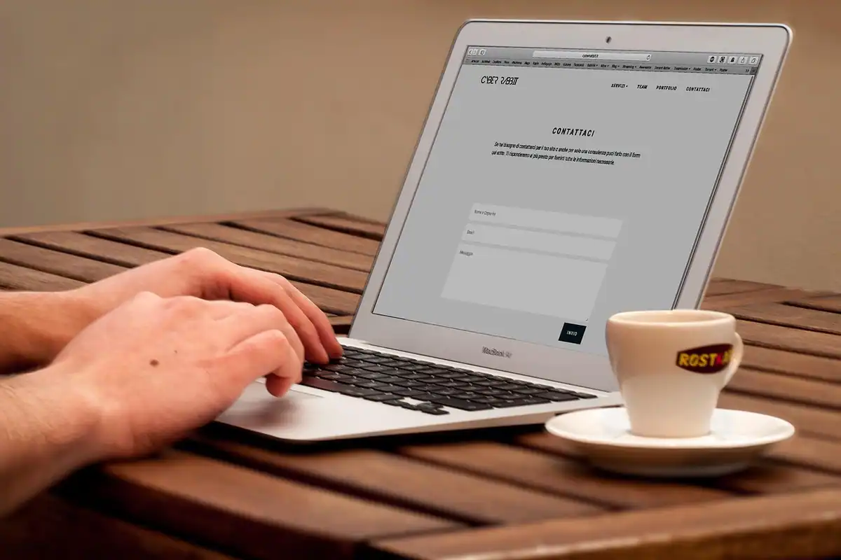 Mâini care tastează pe laptop, lângă o ceașcă de cafea, pe o masă de lemn