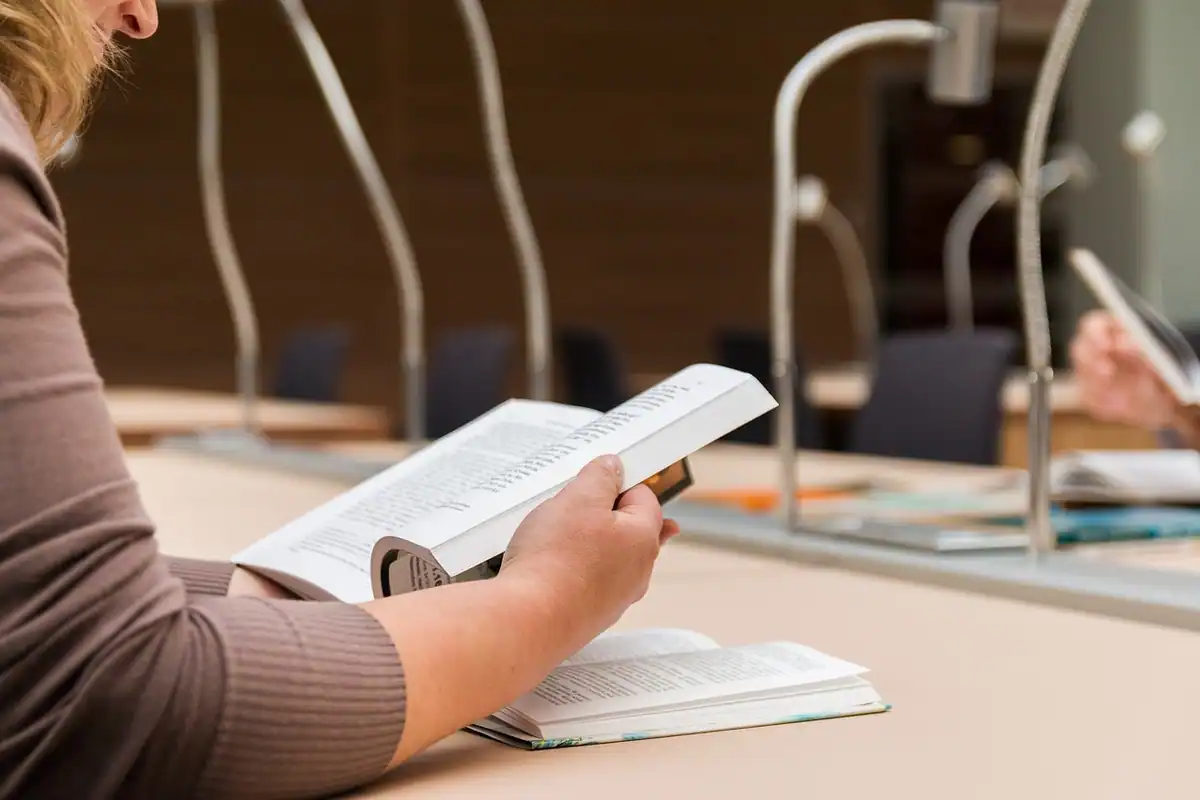 Femeie care citește o carte deschisă la o masă, într-o bibliotecă